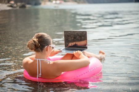 A woman is sitting on a pink inflatable raft in the water, using a laptop. Concept of relaxation and leisure, as the woman is enjoying her time in the water while working on her laptop.の写真素材