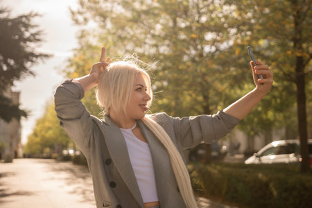 A woman is taking a picture of herself with her cell phone. She is wearing a gray jacket and scarf. The scene is set in a city with trees and cars in the background.の写真素材