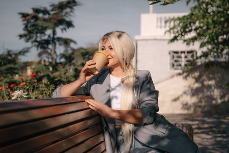 A blonde woman is sitting on a bench with a cup of coffee in her hand. She is smiling and looking out into the distance.の写真素材