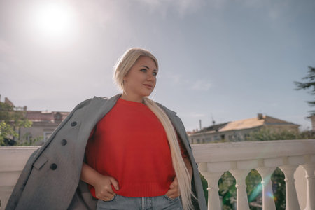 A blonde woman wearing a red shirt and gray jacket stands on a balcony. She is wearing ripped jeans...