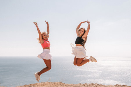 Two women are jumping in the air on a beach. The sky is blue and the ocean is in the background. Scene is joyful and energetic.の写真素材