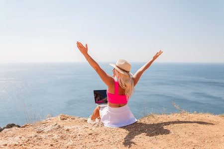 A woman is sitting on a beach with her arms outstretched, wearing a pink top and a straw hat. She is looking out at the ocean, and there is a laptop on the ground in front of her.の写真素材