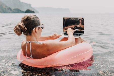 A woman is sitting on a pink inflatable raft in the ocean, reading a laptop. Concept of relaxation...