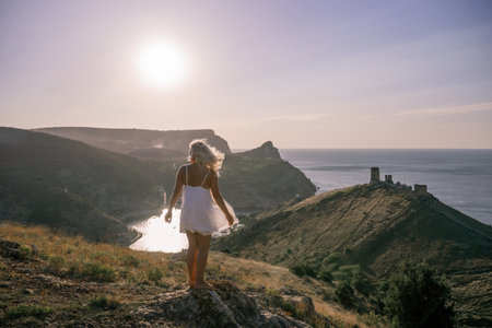 A woman stands on a hill overlooking a body of water. The sky is clear and the sun is shining brightly. The woman is enjoying the view and the peaceful atmosphere.の写真素材