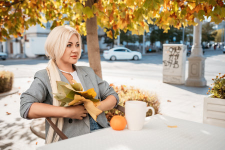 A blonde woman sits at a table with a cup of coffee and a stack of papers. She is wearing a gray jacket and a necklace. The scene is set in a park with trees and benches.の写真素材