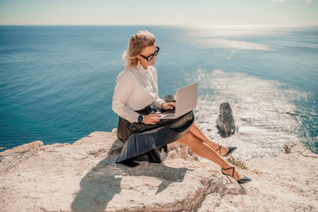 Woman on nature in white shirt and black skirt. She works with a tablet in the open air with a beautiful view of the sea. The concept of remote work.の写真素材