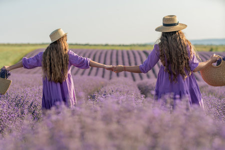 Mom and daughter are running through a lavender field dressed in purple dresses, long hair flowing and wearing hats. The field is full of purple flowers and the sky is clear.の写真素材