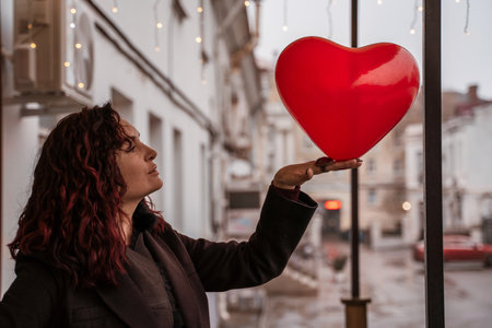 A woman holding a red heart balloon. The woman is wearing a black coat and has red hair. The scene is set in a city street with cars and a building in the background.の写真素材