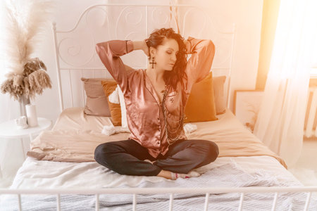 Photo of a middle aged woman in pajamas stretching her arms and smiling while sitting on the bed...