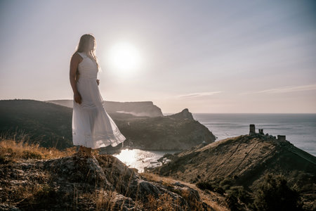 A woman stands on a rocky hill overlooking the ocean. She is wearing a white dress and she is enjoying the view. The scene is serene and peaceful, with the sun shining brightly in the background.の写真素材