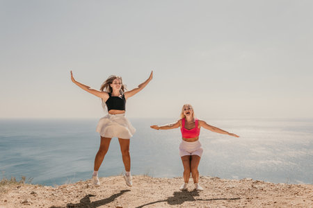 Two women are jumping in the air on a beach. The sky is blue and the ocean is in the background. Scene is joyful and energetic.の写真素材
