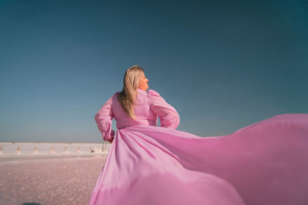 Woman Pink Dress Wind Salt Flats Pose. A woman in a pink dress twirls with her arms above her head, the fabric billowing in the wind. The background is a vast, white, sandy expanse under a blue sky.の写真素材