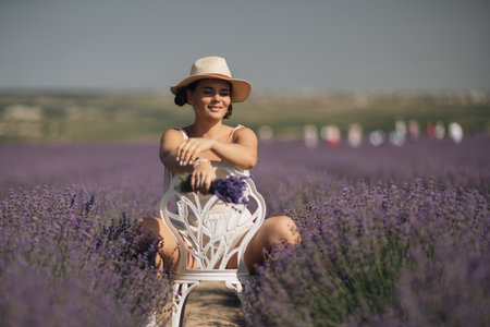 woman sitting in a field of lavender and wearing a straw hat. She is smiling and holding a bouquet of flowers. Scene is peaceful and serene, as the woman is surrounded by the beauty of natureの写真素材