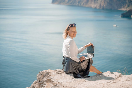Business woman on nature in white shirt and black skirt. She works with an iPad in the open air with a beautiful view of the sea. The concept of remote work.の写真素材