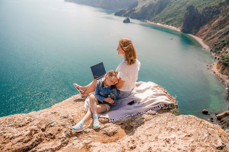 Freelance woman with her daughter working on a laptop by the sea, typing on the keyboard, enjoying the beautiful view, highlighting the idea of remote work.の写真素材