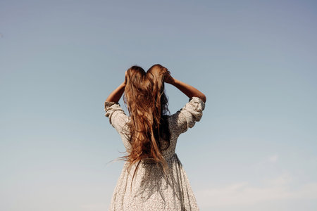 woman with long hair is standing in a field with a blue sky above her. She is wearing a white dress...