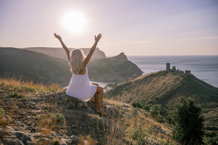 Happy woman is sitting on a hillside, wearing a white dress. She is surrounded by a beautiful landscape, with a body of sea in the background. Concept of peace and happiness.の写真素材