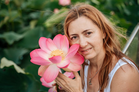 A woman is holding a pink lotus flower and smiling. The flower is surrounded by green leaves, and...