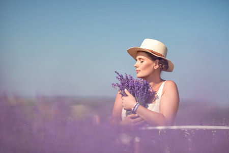woman sitting in a field of lavender and wearing a straw hat. She is smiling and holding a bouquet...