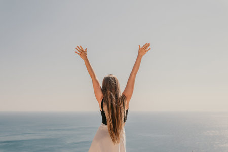 A woman stands on a rocky beach with her arms raised in the air. The sky is clear and the ocean is...