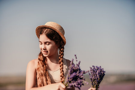 girl is holding a bunch of lavender purple flowers in her hands and wearing a straw hat. She is smiling and she is enjoying herself. The scene is set in a field of lavender, which adds to the peaceful.の写真素材