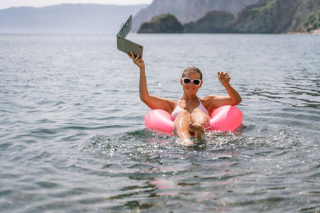 A woman is floating in a pink inflatable raft with a laptop in her hand. She is smiling and she is...
