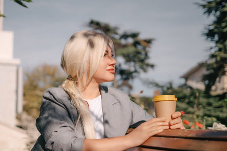 A blonde woman is sitting on a bench with a cup of coffee in her hand. She is smiling and looking...