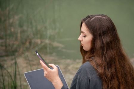 A woman is sitting on the ground with a laptop and a cell phone in her hands. She is using the laptop to browse the internet and the cell phone to communicate with someone.の写真素材