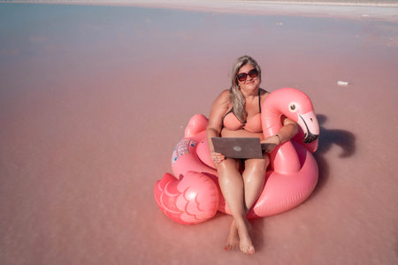Woman Laptop Flamingo Pink Lake Working Remotely. A woman in a bikini sits on a pink flamingo float in a pink lake, working on a laptop.の写真素材
