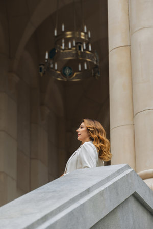 Blonde woman looks upwards at a chandelier while standing on a grand staircase.