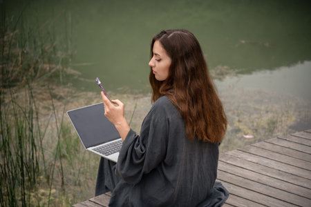 A woman is sitting on the ground with a laptop and a cell phone in her hands. She is using the...