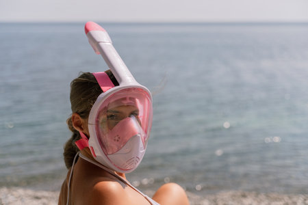 A woman wearing a pink snorkel mask is sitting on a beach. The water is calm and the sky is clear.の写真素材
