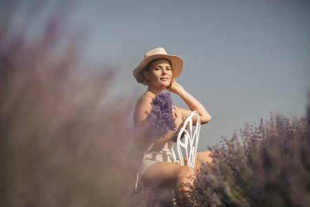 woman sitting in a field of lavender and wearing a straw hat. She is smiling and holding a bouquet of flowers. Scene is peaceful and serene, as the woman is surrounded by the beauty of natureの写真素材