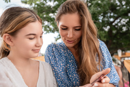 Woman Teenager Smartphone Outdoors: Mother assists daughter with smartphone outdoors, likely teaching or sharing information. Summer daytime.の写真素材