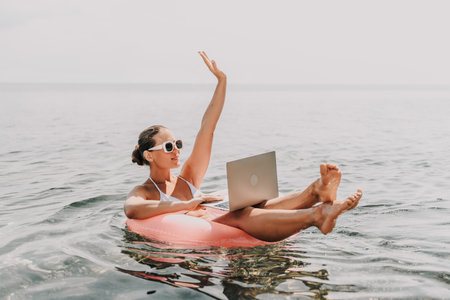 A woman is sitting on a pink inflatable raft in the ocean, holding a laptop. She is smiling and she is enjoying her time. Concept of relaxation and leisure.の写真素材
