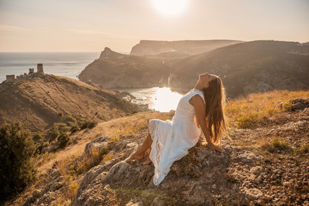 A woman in a white dress is sitting on a rock overlooking a body of water. She is enjoying the view and taking in the scenery.の写真素材