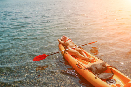 A woman is laying on a kayak in the water. The kayak is orange and has a paddle on it. The woman is wearing a red hat.の写真素材