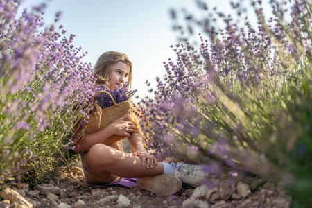 Girl is sitting in a field of purple flowers. She is holding a basket of flowers and smiling. Scene is peaceful and serene, as the girl is surrounded by the beauty of nature.の写真素材