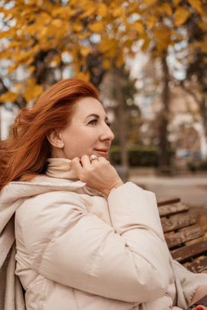 Woman Autumn Park Bench: Redhead relaxes outdoors, enjoying fall foliage, serene autumnal scene.の写真素材