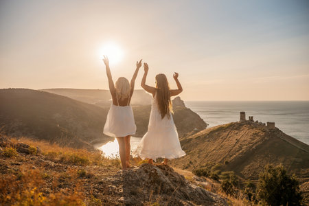 Two women are standing on a hill overlooking the ocean. They are holding hands and looking out at the water. The scene is peaceful and serene, with the sun shining brightly in the background.の写真素材