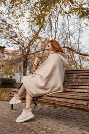 Woman Park Bench Autumn: Cozy hooded beige poncho, enjoying coffee, fall outdoor relaxation.の写真素材