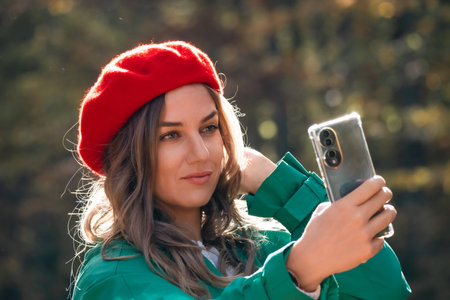 Woman Selfie Smartphone Autumn Outdoors: Red Beret, Green Jacket, Fall Sunlight.の写真素材
