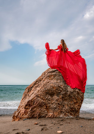 woman sea red dress. Woman with long hair on a sunny seashore in a red flowing dress, back view, silk fabric waving in the wind. Against the backdrop of the blue sky and mountains on the seashore.の写真素材