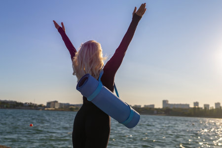 Woman Yoga Mat Beach Sunset: A woman in black sportswear raises her arms in celebration while standing on a beach with her yoga mat. The sun is setting behind her, casting a golden glow over the water.の写真素材