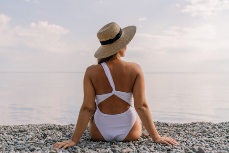 A woman wearing a white swimsuit and a straw hat is sitting on a beach. The beach is rocky and the sky is cloudy.の写真素材