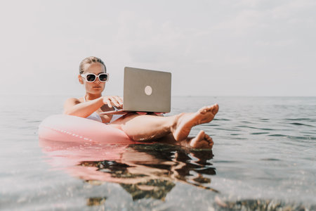 A woman is sitting on a pink inflatable raft in the ocean, using a laptop. Concept of relaxation and leisure, as the woman is enjoying her time in the water while working on her laptop.の写真素材