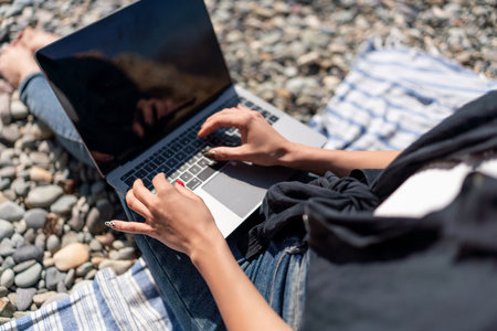 Laptop Beach RemoteWork: Woman types outside on pebble beach, bright day for leisure.の写真素材