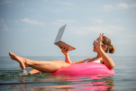 A woman is floating on a pink inflatable raft in the ocean while holding a laptop. Concept of relaxation and leisure, as the woman is enjoying her time in the water while working on her laptop.の写真素材