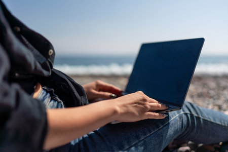 Laptop Beach Work: Woman typing on laptop near sea, midday, for remote work leisure on sunny rocky shore.の写真素材
