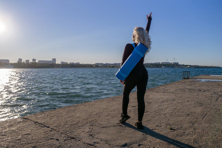 Woman Yoga Mat Beach - A woman in black leggings holds a blue yoga mat on her back while standing on a concrete pier overlooking a body of water. She raises her hand in the air. The sky is blue and the sun is shining. She appears to be enjoying the view and the beautiful weather.の写真素材